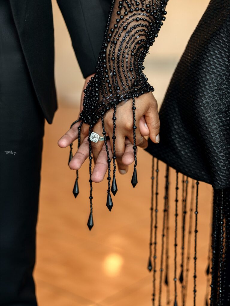 Close-up of two hands interlocked, showcasing intricate black beaded sleeve details and a diamond ring.