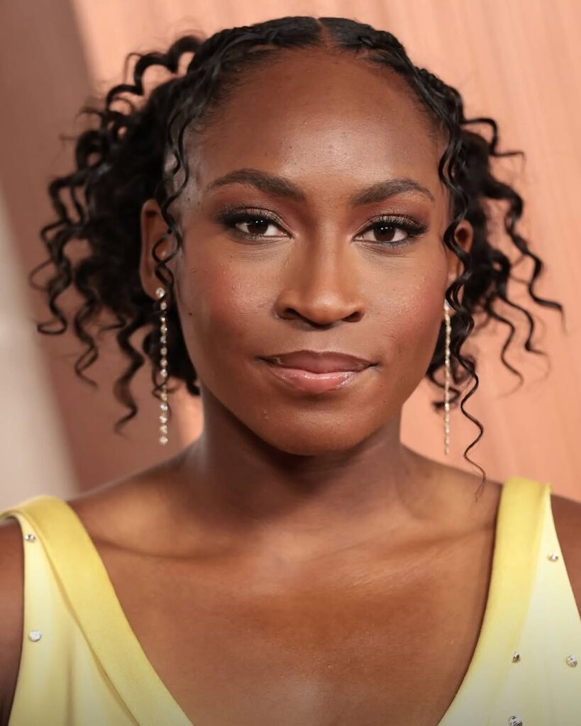 A poised woman with curly hair and sparkling earrings, wearing a yellow dress, smiles softly against a warm background.