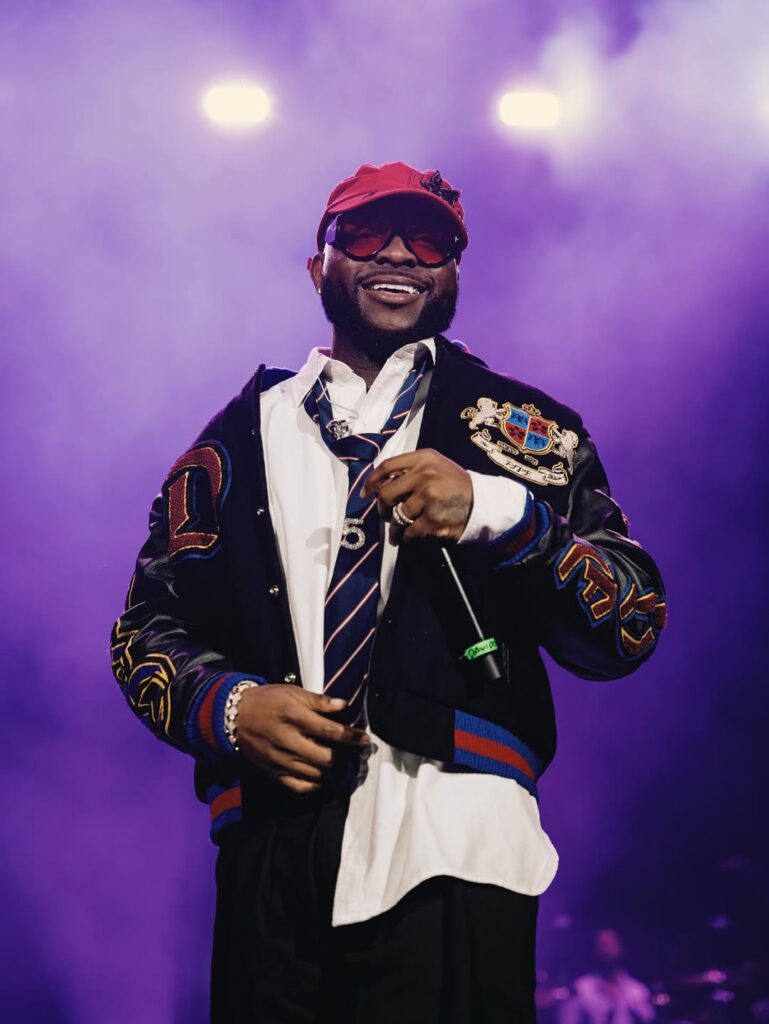 An artist wearing a stylish outfit and a red cap smiles while performing on stage under colorful lights.