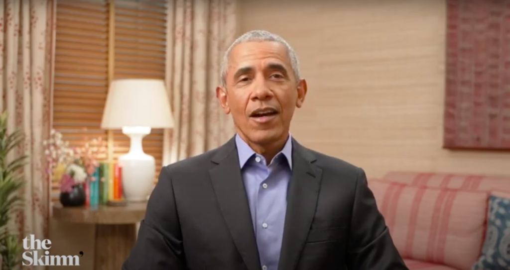 A seated individual in a dark suit with a light blue shirt, smiling and speaking, in a cozy indoor setting with decorative elements like a lamp and flowers.